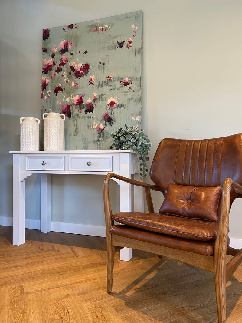 Full view of the white painted dressing table holding two textured ceramic vases below a leaning floral canvas, with a tan leather and oak armchair in front on herringbone wood flooring