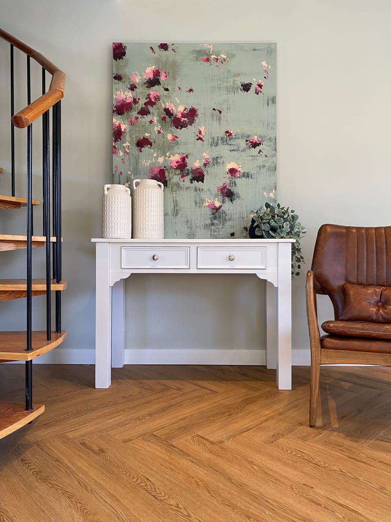 View of the dressing table beside an open oak and black metal staircase, its two drawers fitted with chrome knobs and a sage and pink abstract canvas leaning behind the painted top