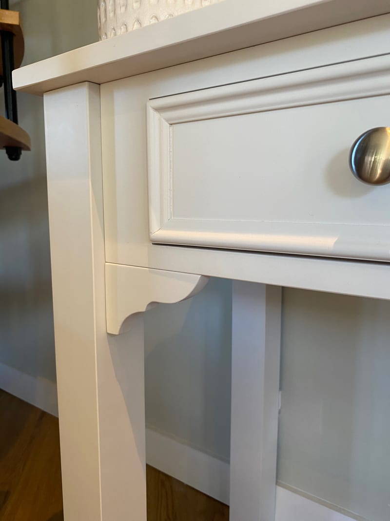 Close view of the corner of the white painted dressing table, showing the square leg, the shaped bracket beneath the apron and the moulded edge of the drawer front above