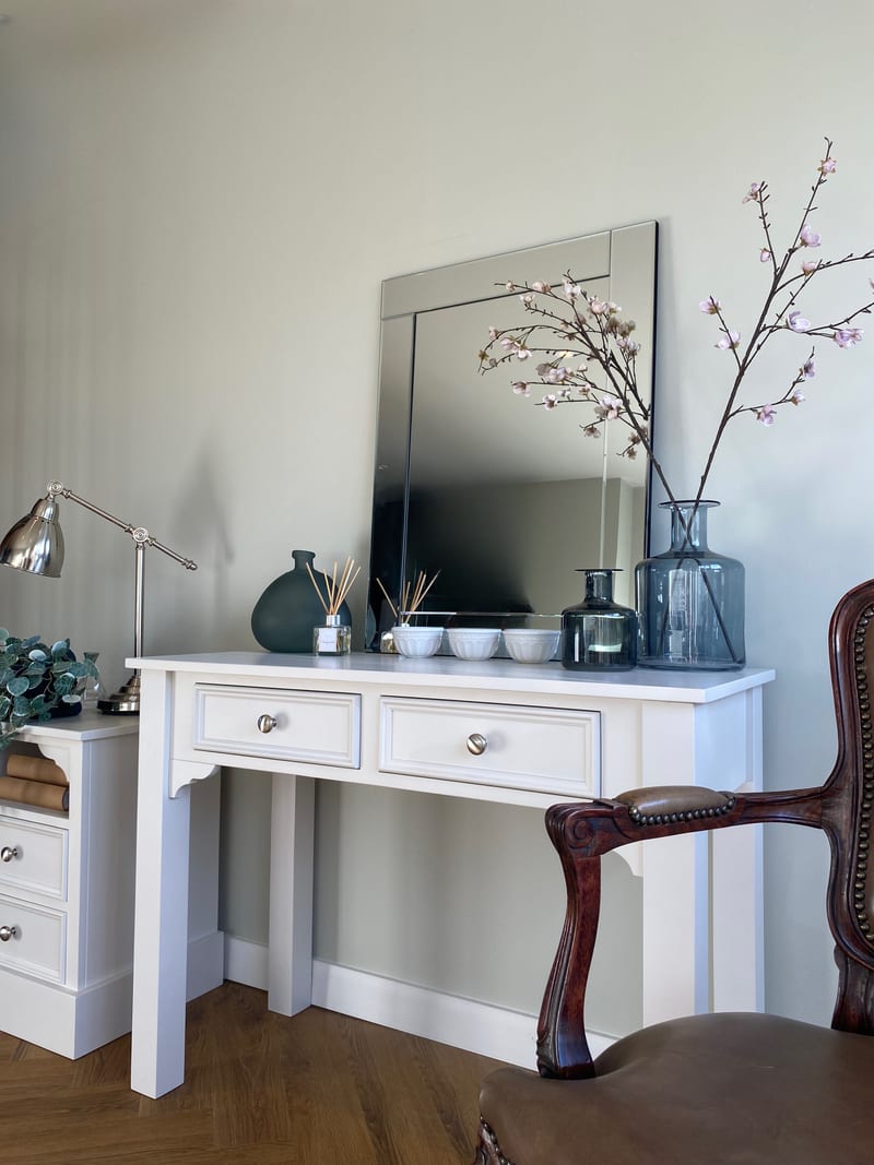 Three-quarter view of the dressing table beneath a bevelled mirror, with a smoked glass bottle of blossom, a dark round vase and three small white bowls on the painted top