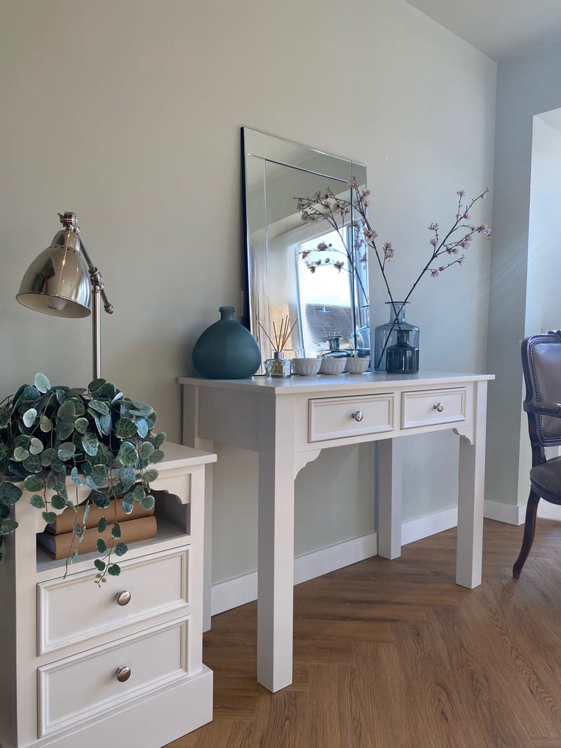 Angled view of the dressing table from the side, showing the shaped brackets and square legs, with a mirror, teal vase and blossom branches on top and a trailing plant on the cabinet alongside
