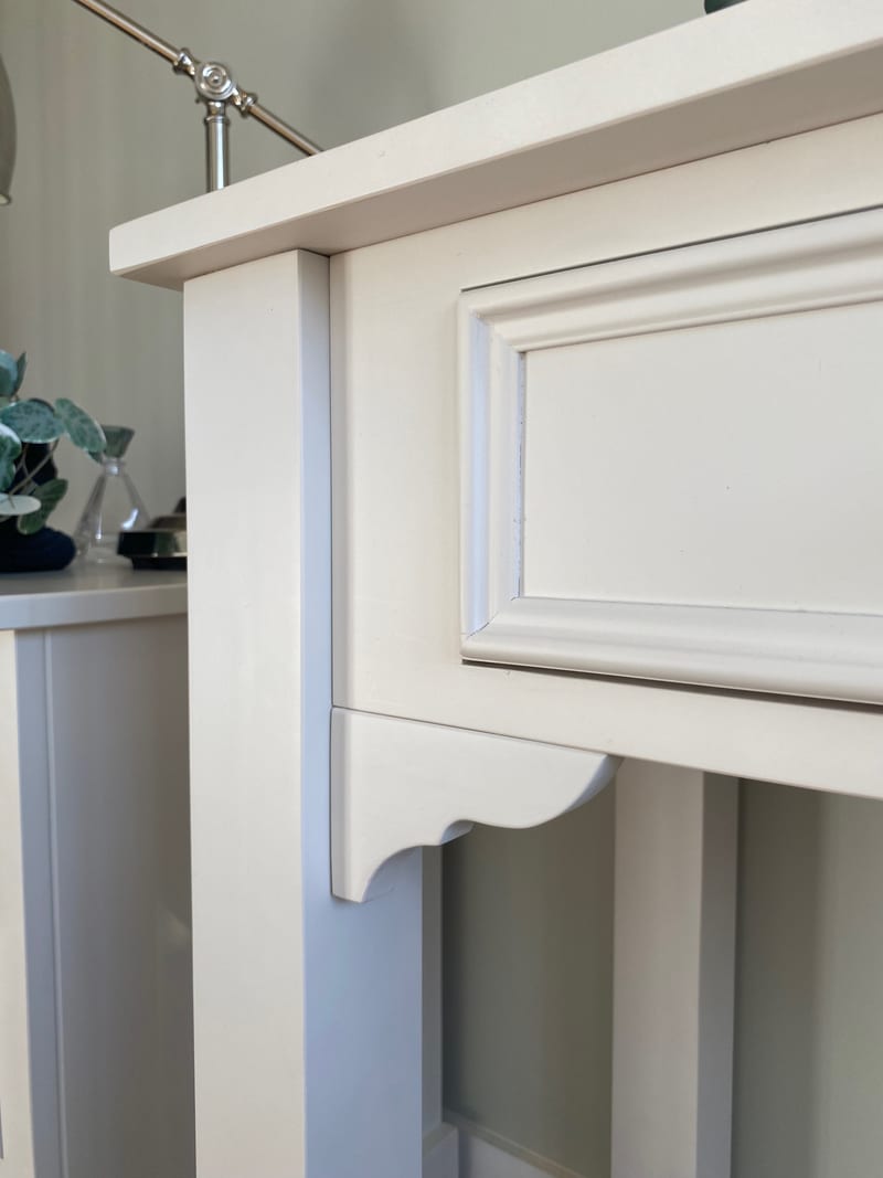 Detail of the top overhang and drawer front moulding on the painted dressing table, with the scrolled bracket and square leg below and a glimpse of the bedside cabinet behind