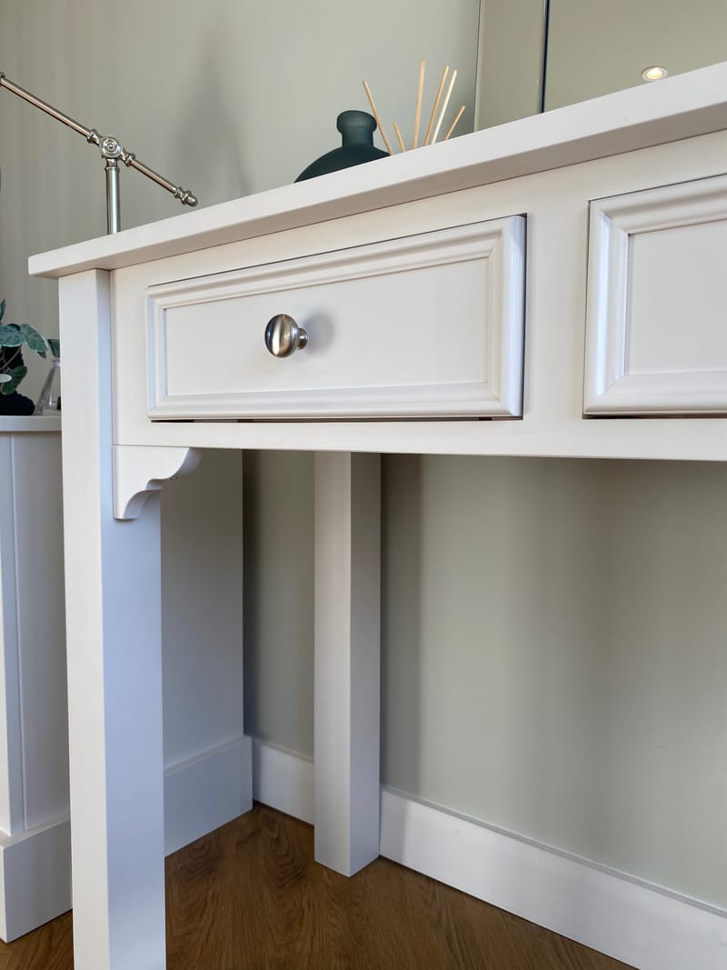 Close view of one moulded drawer front and its round chrome knob on the painted dressing table, with the shaped bracket, square legs and the open kneehole space beneath