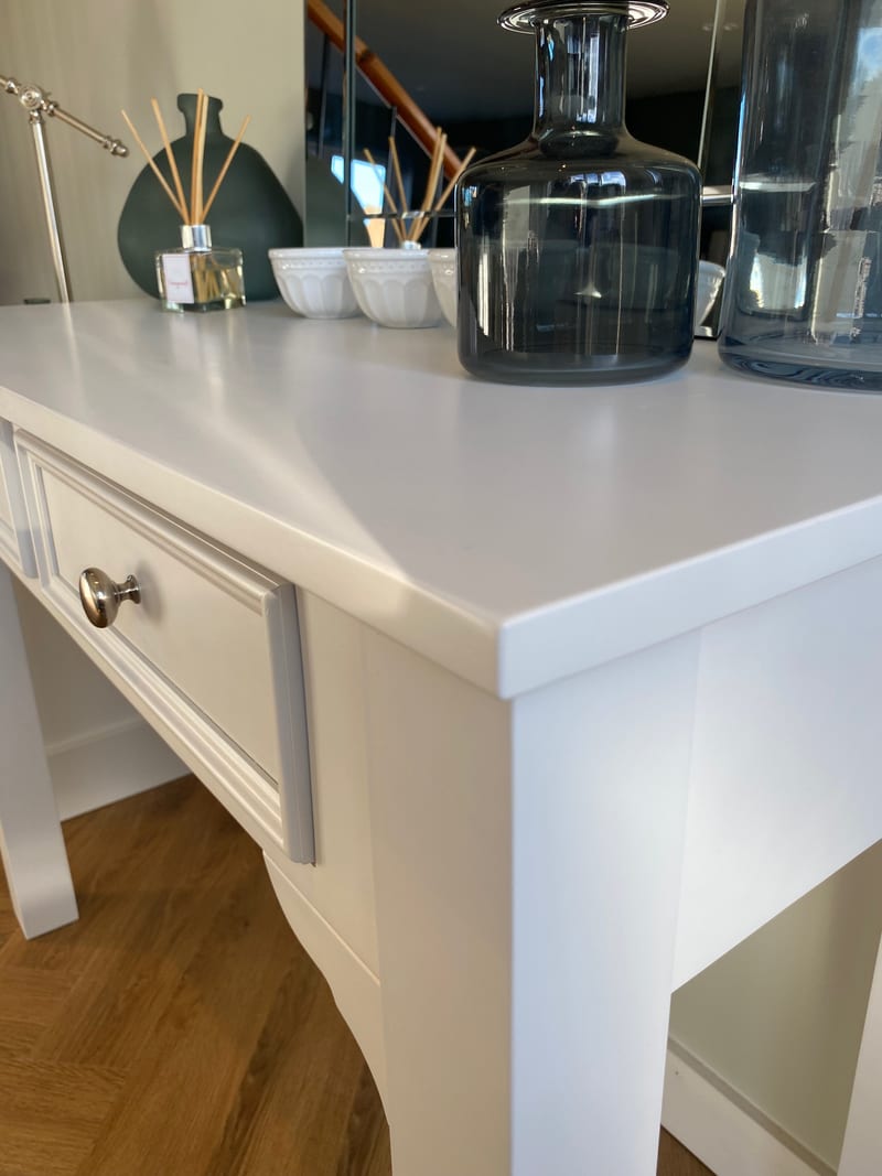 Close view along the painted top of the dressing table, showing the square edge and corner beside smoked glass bottles, white bowls and a reed diffuser with the mirror behind