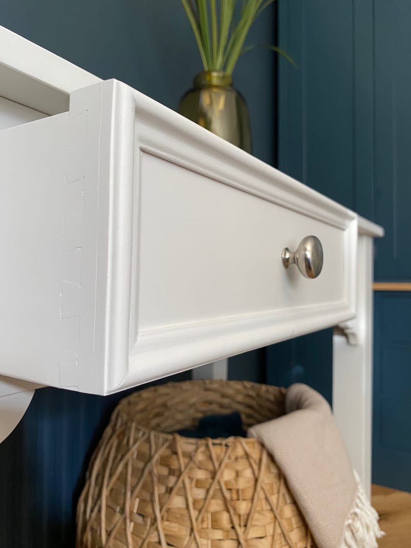 Close view of a drawer pulled out from the cream painted dressing table, showing the moulded front, round chrome knob and the painted drawer side, with a woven basket below