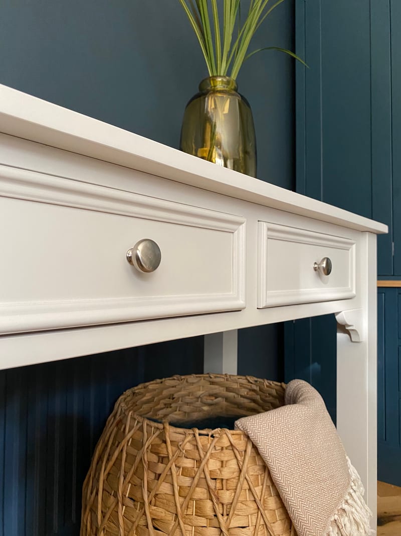 Angled close view of the two moulded drawer fronts and the square top edge, with brushed nickel knobs, an olive glass vase on the table and a fringed throw over the basket beneath