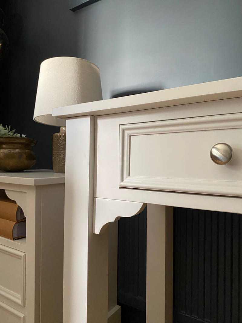 Corner detail of the cream painted dressing table showing the moulded drawer front and satin nickel knob, the shaped bracket and leg, with a lamp and brass pot on the cabinet behind
