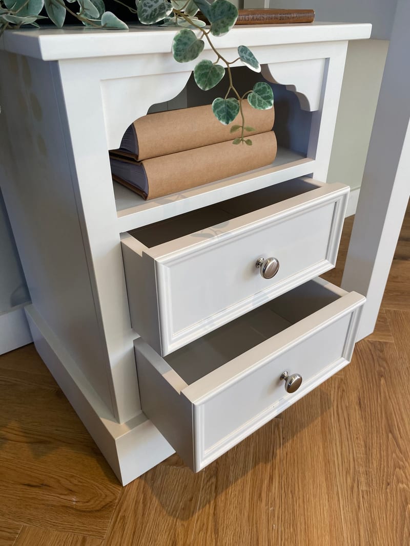 Angled view from above of the white bedside cabinet with both drawers pulled open, showing the painted drawer boxes, the open shelf above holding two kraft-covered books and the herringbone oak floor