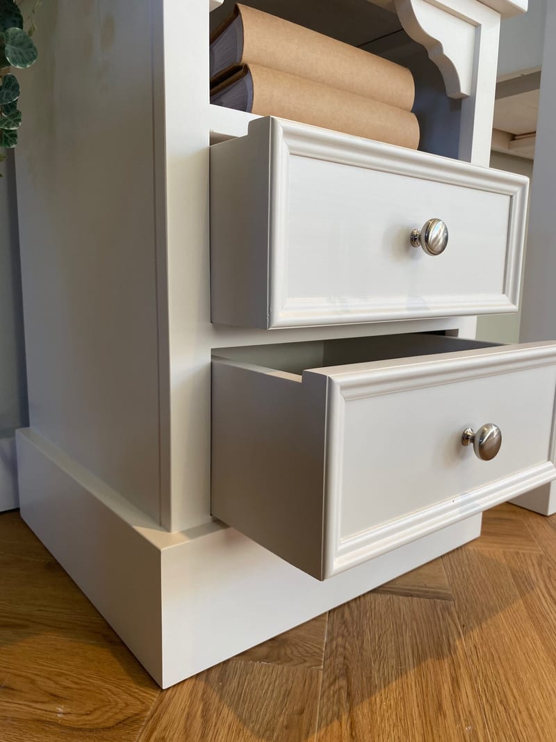 Low close view of the corner of the white painted bedside cabinet with both drawers open, showing the moulded drawer fronts, brushed nickel knobs and the deep plinth base above a herringbone oak floor
