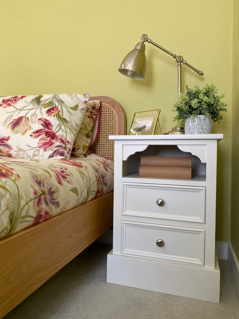 Full view of the white bedside cabinet beside an oak bed with a rattan headboard and floral bedding, its shelf and two drawers set against a yellow-green wall below a brass lamp and framed print