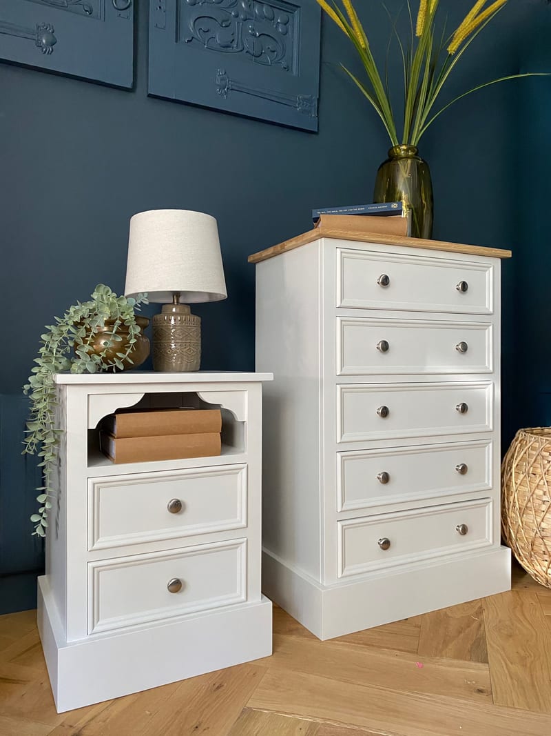 Full view of the white bedside cabinet standing beside a taller five-drawer chest with an oak top against a dark blue panelled wall, both with round metal knobs, on a herringbone oak floor
