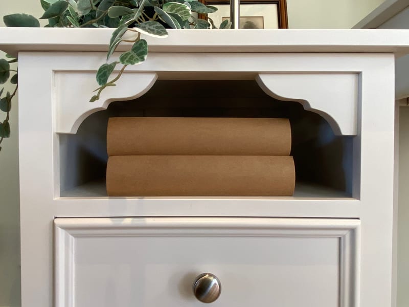 Close view of the open shelf on the white bedside cabinet, showing the shaped scalloped brackets, the painted back panel, two kraft-covered books inside and the knob on the drawer below
