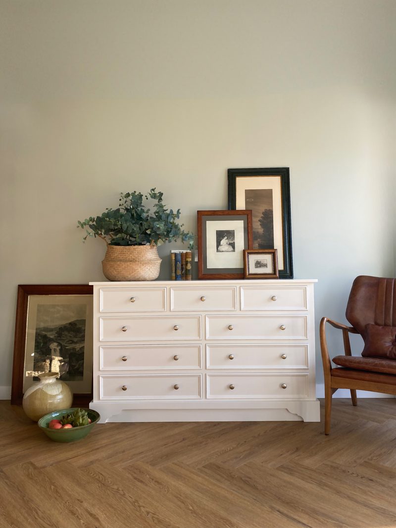 Full view of the nine-drawer painted chest against a pale green wall, styled with a woven basket of eucalyptus, layered framed prints and books, beside a tan leather armchair on herringbone flooring