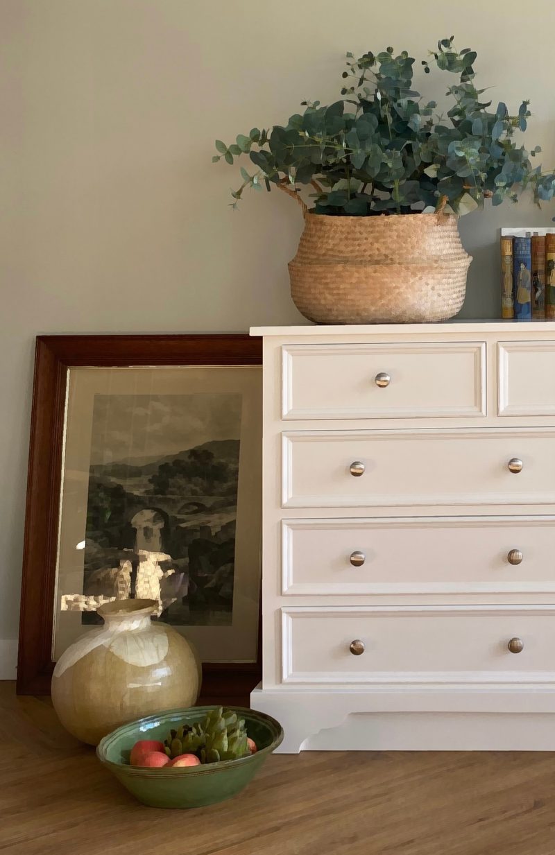 Close view of the left-hand end of the white painted chest, showing four drawer fronts with round metal knobs and the shaped plinth beside a leaning print, olive glazed vase and bowl of apples