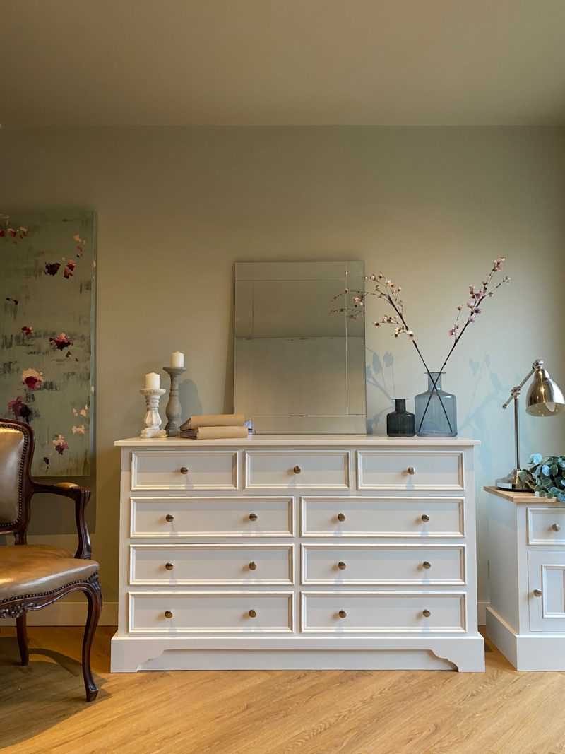 Full view of the nine-drawer painted chest in warm evening light, topped with a leaning frameless mirror, stone candlesticks and blossom stems in glass vases, beside a carved wooden armchair