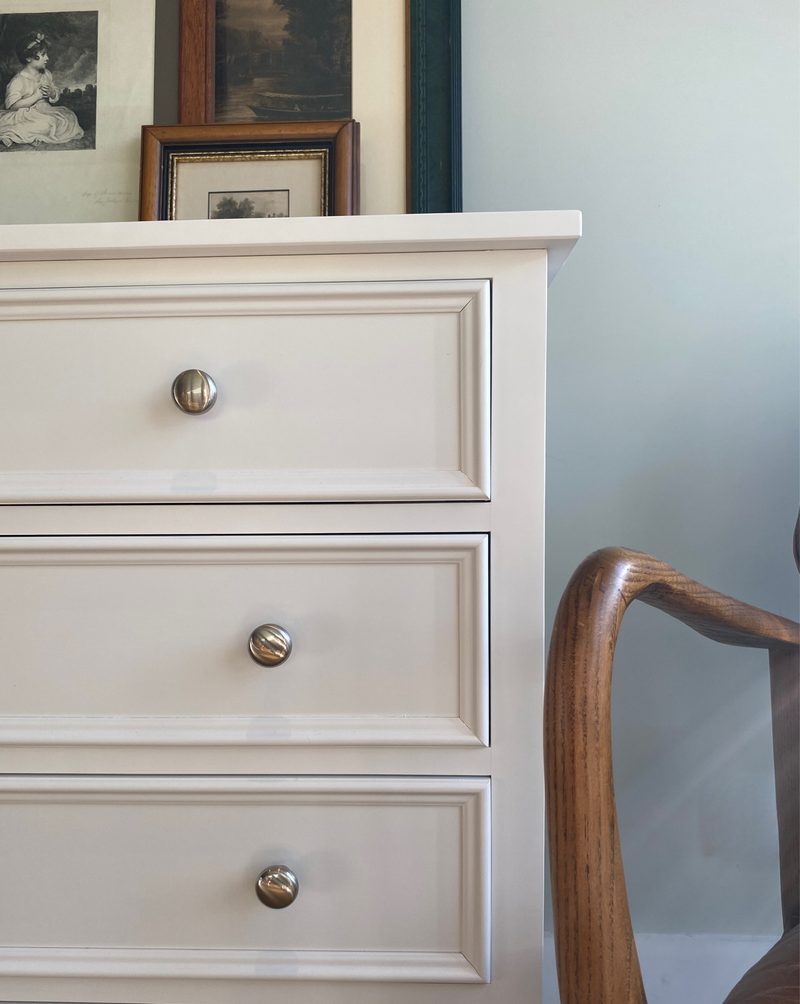 Close view of the top corner of the painted chest, showing the moulded top edge and three panelled drawer fronts with round metal knobs, framed prints above and a wooden chair arm alongside