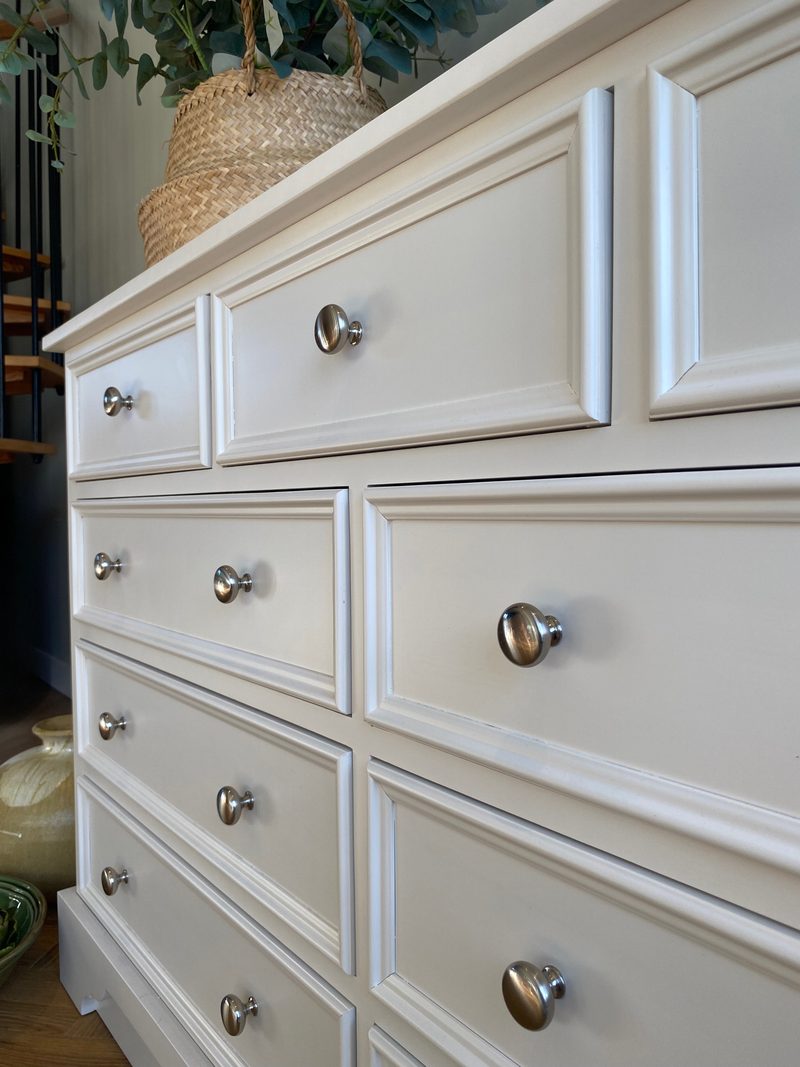 Sharply angled view along the front of the painted chest, showing the panel mouldings and rows of round metal knobs receding towards the seagrass basket of eucalyptus on the top