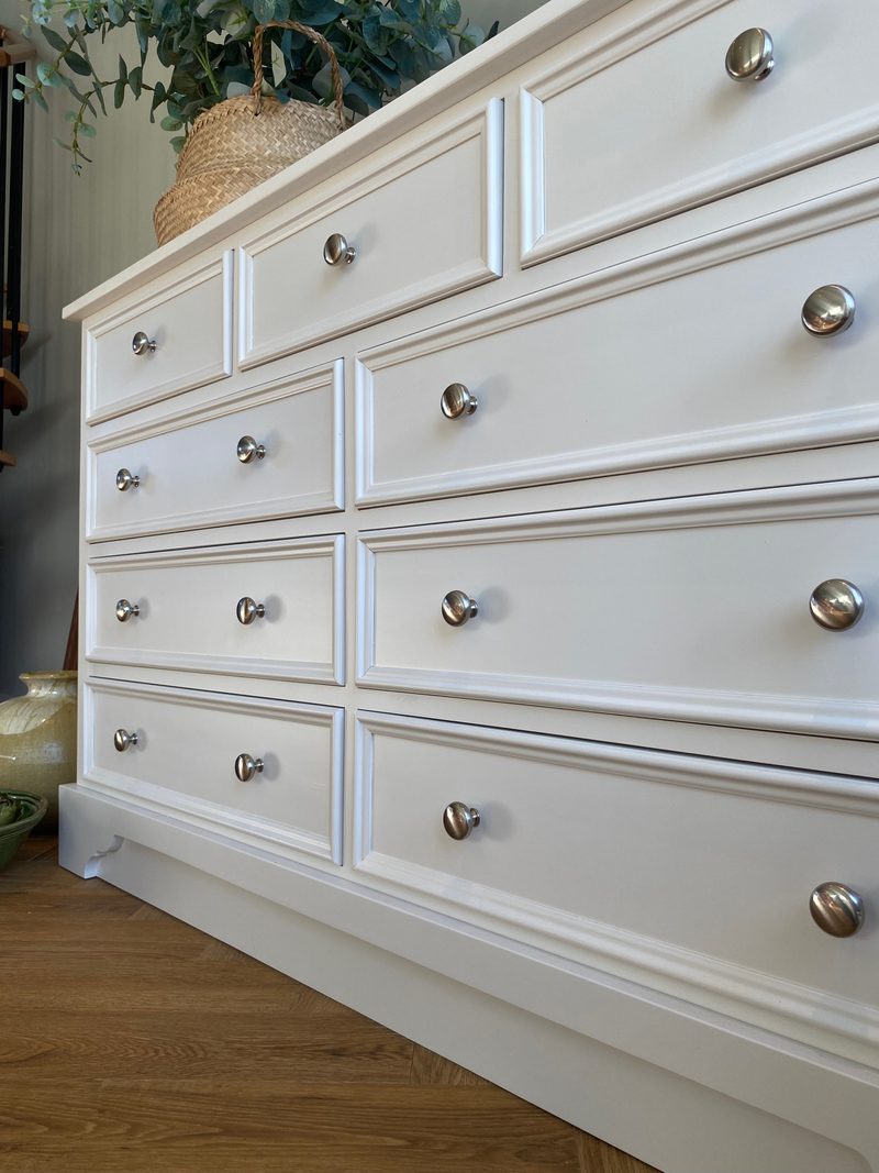 Angled view of the white painted chest showing three short drawers above three pairs of long drawers, each long front carrying two round metal knobs, above the shaped plinth on an oak floor