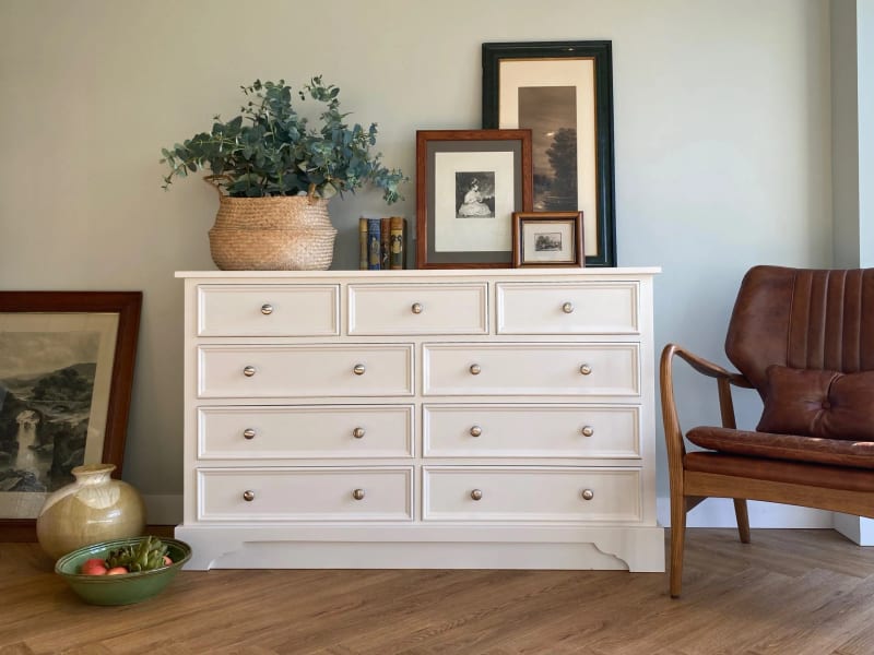 Cream painted Carlton chest with three short drawers above three pairs of long drawers on a shaped plinth, styled with eucalyptus, books and framed prints beside a tan leather chair