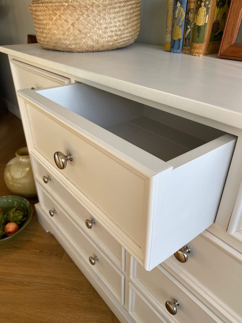 Detail of one long drawer pulled fully out, showing the painted drawer box, its planked base and the polished metal knob, with a woven basket and old books resting on the top above