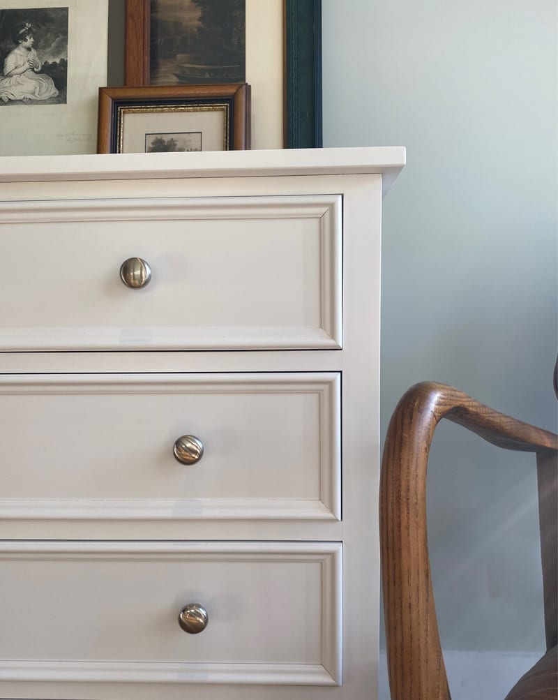 Close view of the top corner of the white painted chest of drawers, showing the moulded top edge, three panelled drawer fronts with round metal knobs and framed prints leaning above