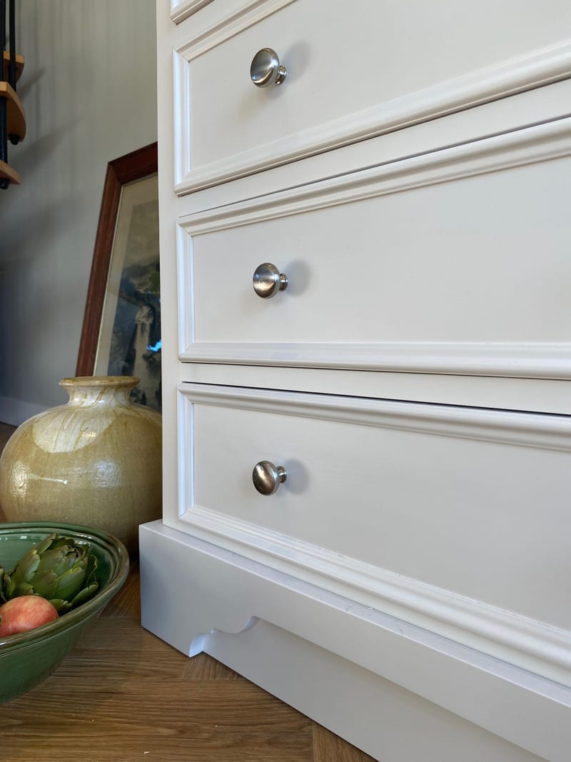 Close view of the lower drawer fronts of the painted chest and the moulded plinth with its shaped cut-out, beside a large glazed ochre vase and a green bowl of artichokes on the oak floor