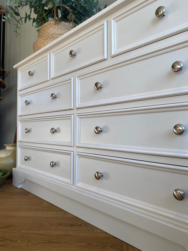 Angled view of the painted chest of drawers showing three short drawers above three pairs of deeper drawers, all with round metal knobs, above a moulded plinth on an oak floor