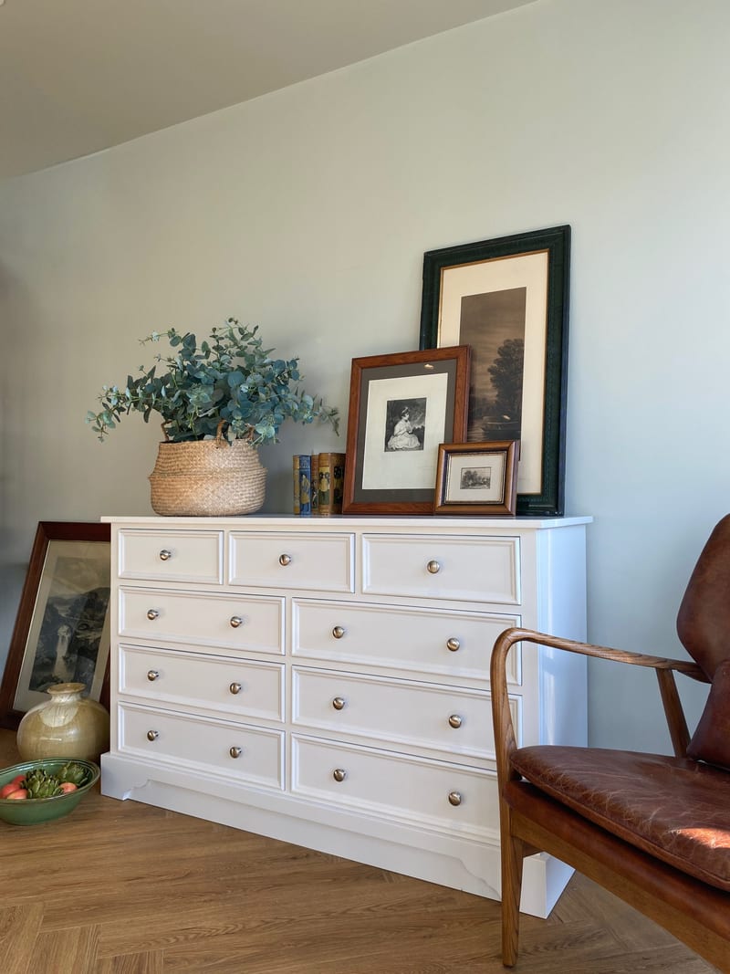 Angled full view of the white painted chest of drawers with framed prints and a basket of eucalyptus on top, its nine drawers and plinth base seen from the left beside a leather armchair