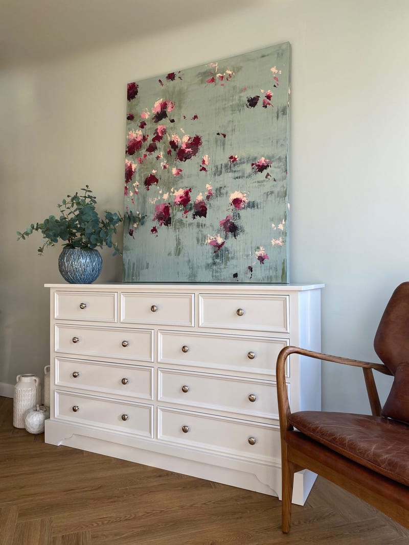 Full view of the white painted chest of drawers beneath a large abstract floral canvas, with a blue textured vase of eucalyptus on top and pale ribbed vases on the floor alongside