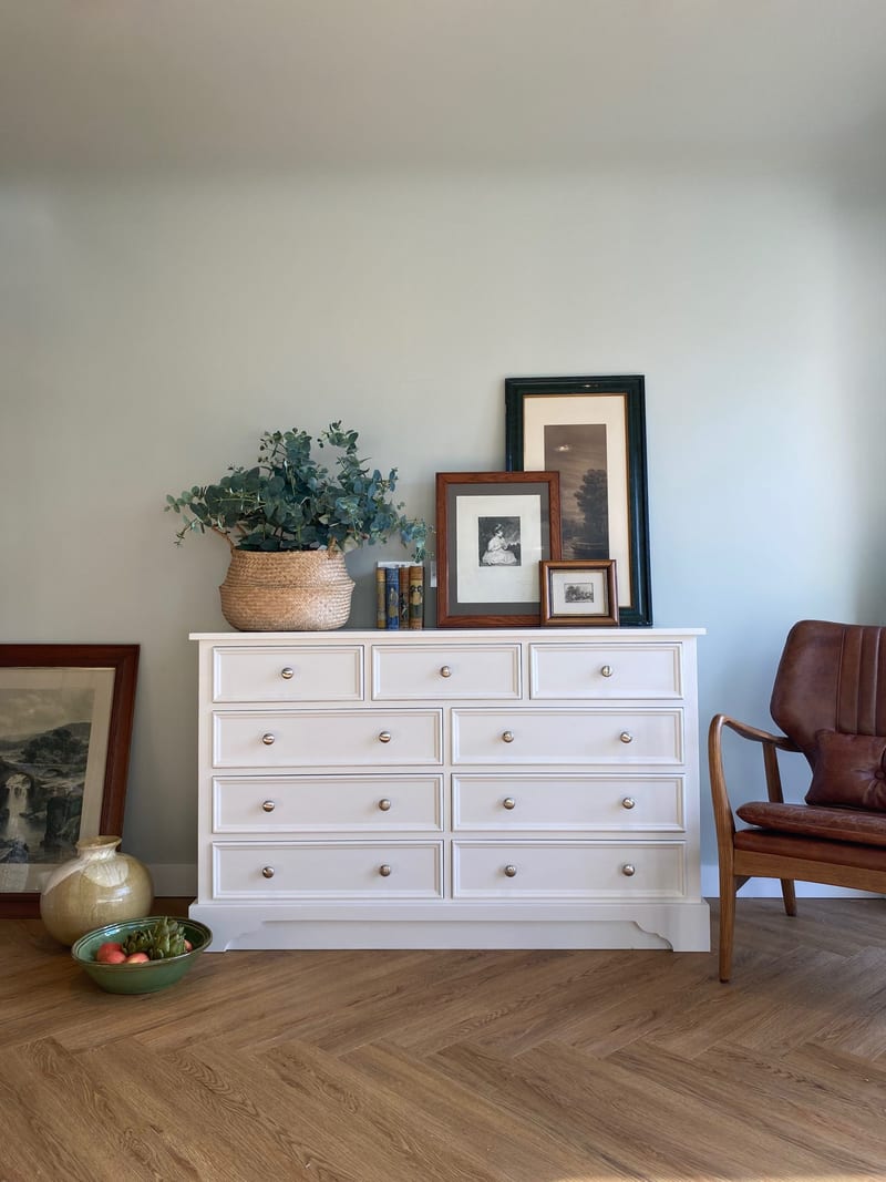 Wide view of the white painted chest of drawers centred on a pale green wall, its three short and six long drawers below a basket of eucalyptus, framed prints and stacked books