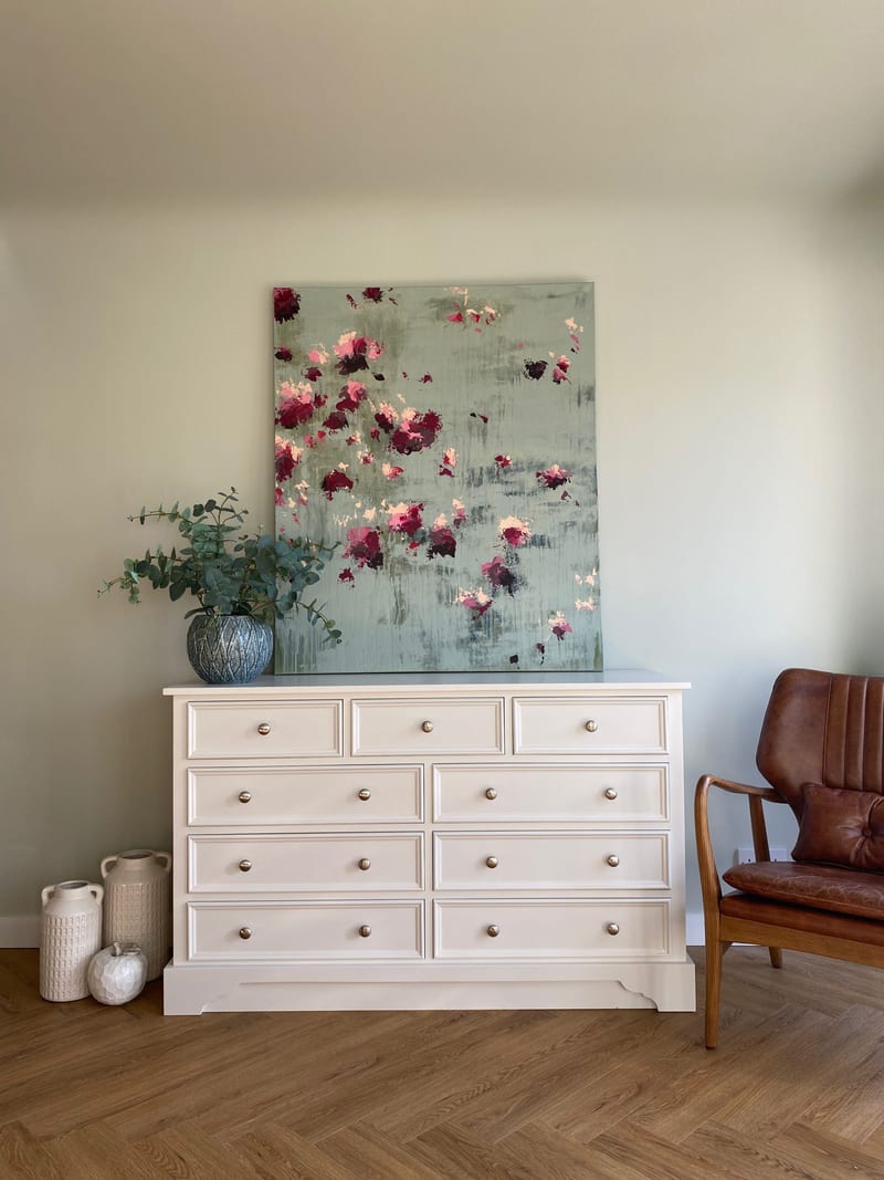 Straight-on view of the white painted chest of drawers below a green and pink abstract canvas, with eucalyptus in a blue vase on top and two ridged cream jars and a small white pot on the floor