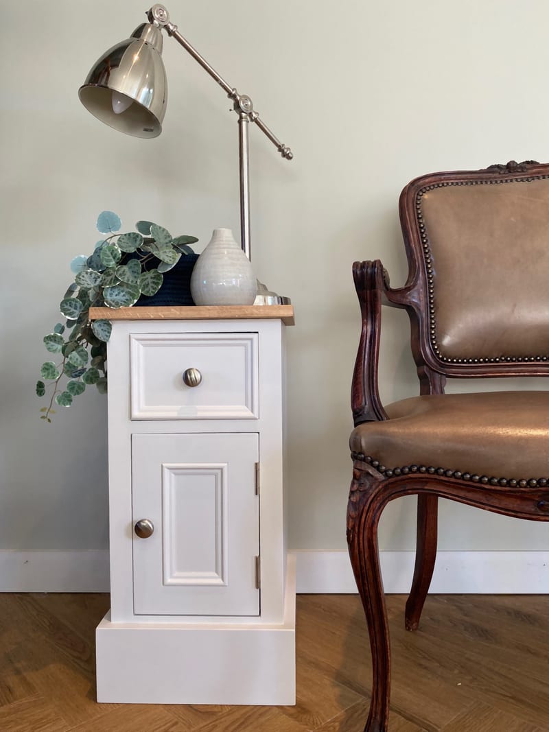 Cream painted Carlton small bedside cabinet with a solid oak top, one drawer above a panelled cupboard door, styled with a vase and trailing plant beside a leather armchair