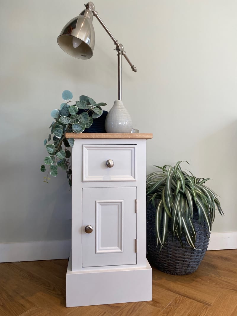 Full length view of the off-white painted bedside cabinet, one drawer above a panelled door with round nickel knobs, its oak top holding a nickel lamp and cream vase beside a spider plant