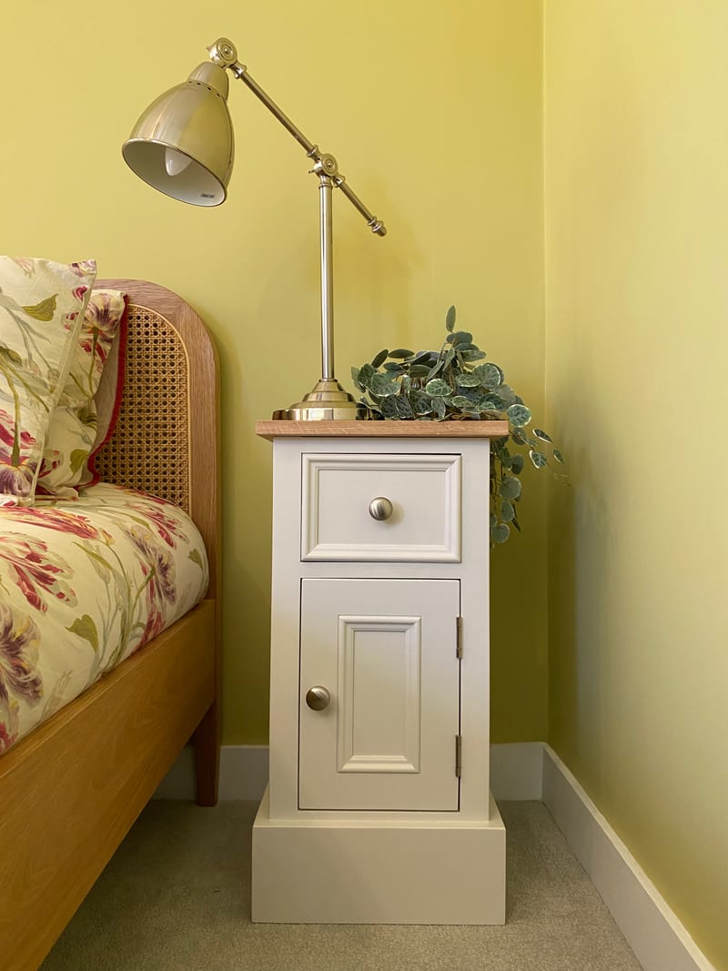 Cream painted bedside cabinet in the corner of a yellow bedroom, its oak top holding a cream shaded task lamp and a trailing faux plant, next to a cane and oak bed with floral bedding