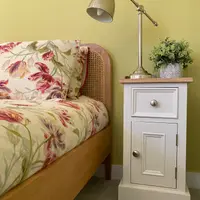 Wider view of the same yellow bedroom, with the cream cabinet beside the floral dressed bed and a small faux plant in a patterned grey pot standing on its oak top beside the lamp