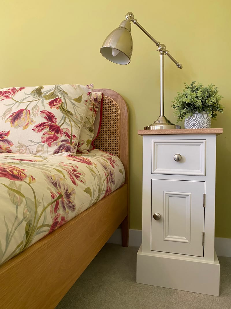 Wider view of the same yellow bedroom, with the cream cabinet beside the floral dressed bed and a small faux plant in a patterned grey pot standing on its oak top beside the lamp