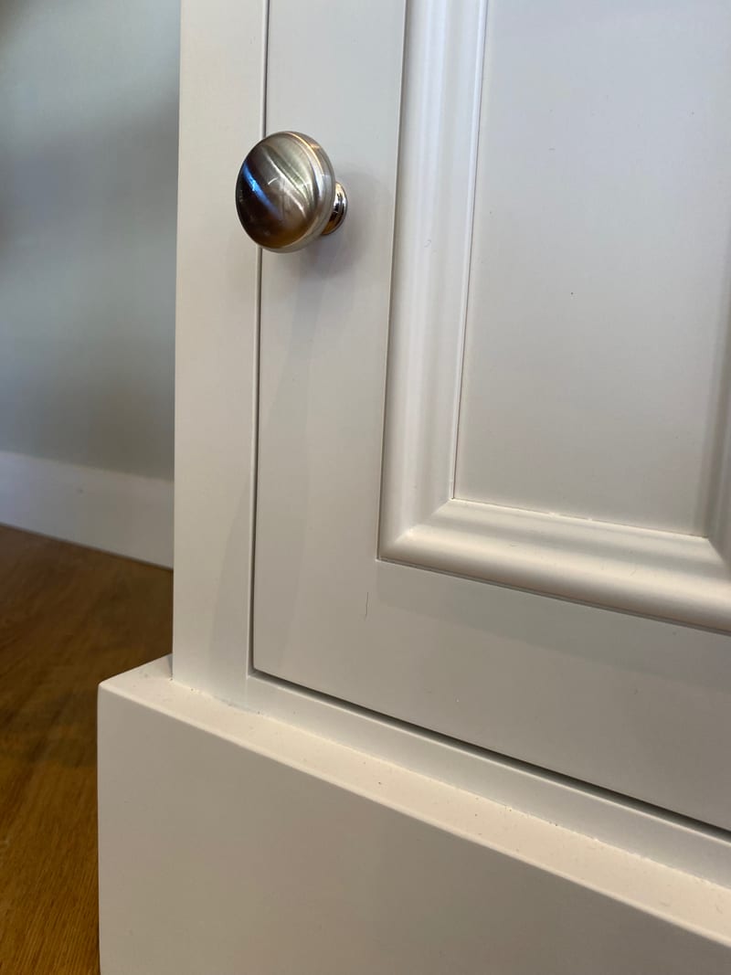 Detail of the panelled door and its round brushed nickel knob, with the moulded frame and the top edge of the painted plinth catching the light against a pale grey wall