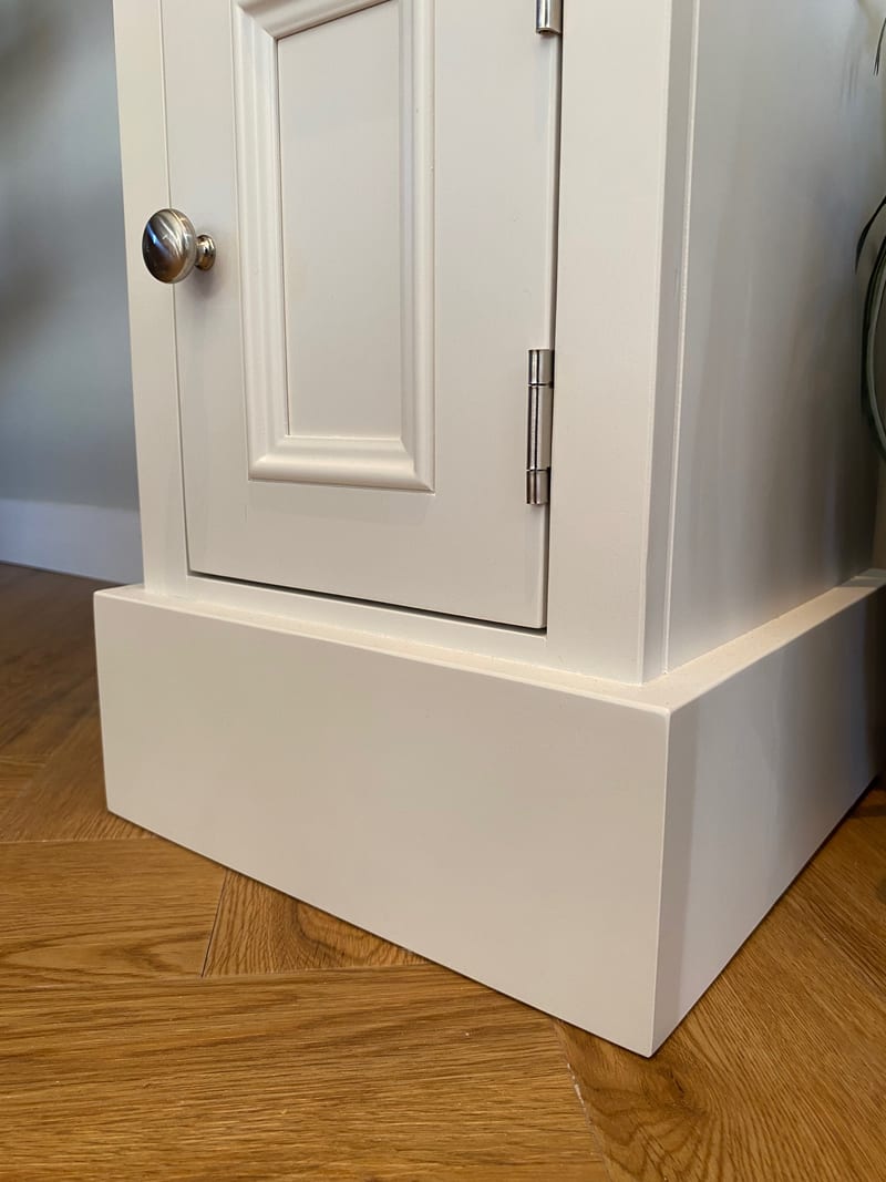 Corner view of the base of the cabinet, showing the deep painted plinth, the moulded door panel and two brushed steel hinges above a herringbone oak floor