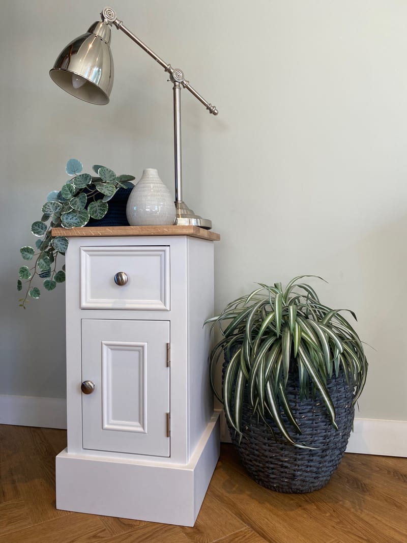 Three-quarter view showing the painted side panel and oak top of the cabinet, the drawer and door fronts with round nickel knobs, and a spider plant in a dark basket alongside