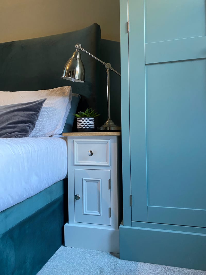 Pale painted bedside cabinet with an oak top tucked between a dark green velvet bed and a teal painted wardrobe, holding a chrome task lamp and a plant in a patterned pot