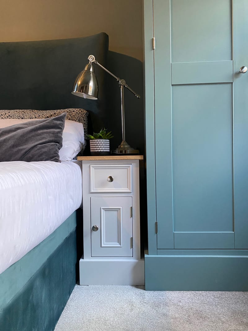 Wider bedroom view of the small cabinet beside a velvet upholstered bed, showing its single drawer, panelled door and plinth, with the teal wardrobe and grey carpet alongside