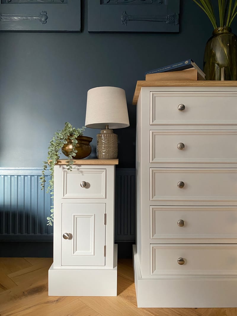 The cabinet and a matching five drawer chest against a dark blue panelled wall, with a table lamp, a brass pot and trailing eucalyptus on the oak top above a herringbone floor