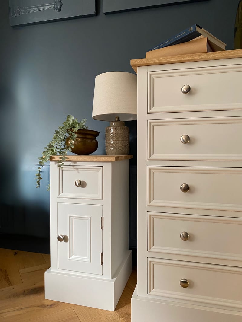 Closer view of the cabinet alongside the taller five drawer chest, both with oak tops and round nickel knobs, styled with a linen shade lamp, a brass pot and stacked books