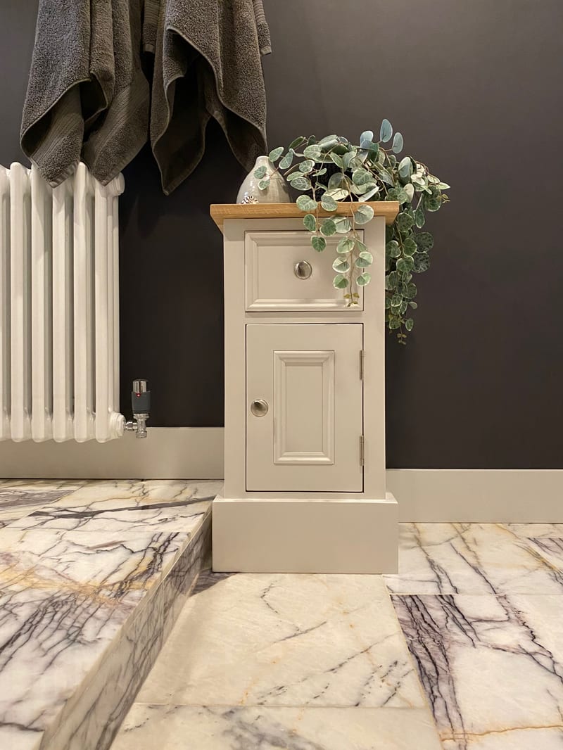 The cabinet used in a bathroom, standing on veined marble tiles against a dark brown wall beside a white column radiator, with towels hanging above and a plant on the oak top