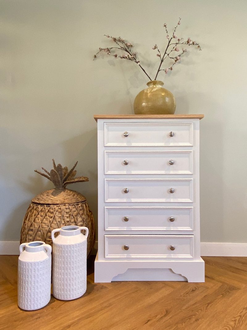Cream painted Carlton tall chest of five deep drawers with a solid oak top and a shaped plinth, styled with a glazed vase of blossom beside woven baskets and ribbed jars