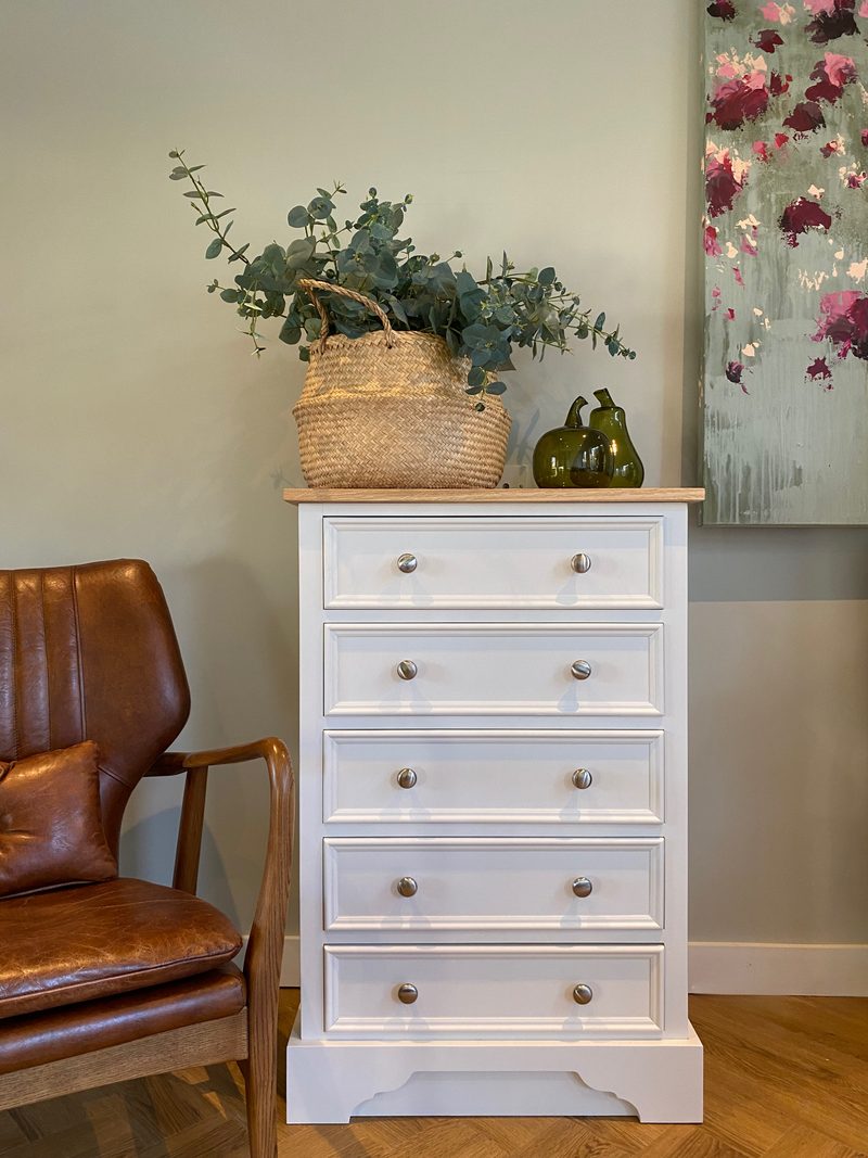Full front view of the white painted five drawer chest with an oak top, ten round nickel knobs and a shaped plinth, styled with eucalyptus in a seagrass basket beside a leather chair