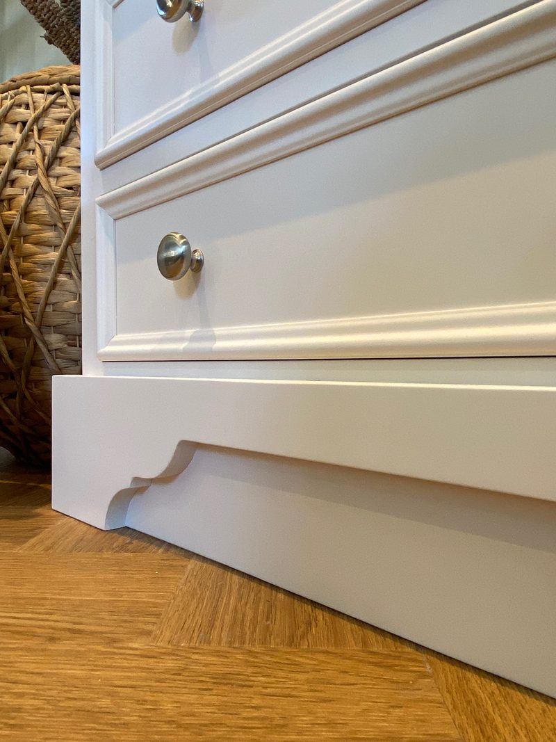 Detail of the lower drawers and the shaped bracket plinth, showing the moulded drawer fronts, two brushed nickel knobs and a woven basket alongside on herringbone oak flooring