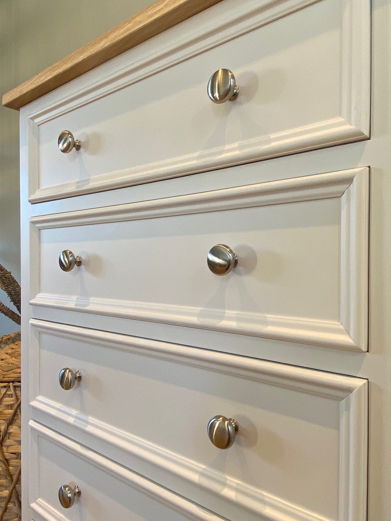 Close angled view along the drawer fronts, showing the bevelled mouldings, the pairs of brushed nickel knobs and the sawn edge of the oak top, with a woven basket just in shot
