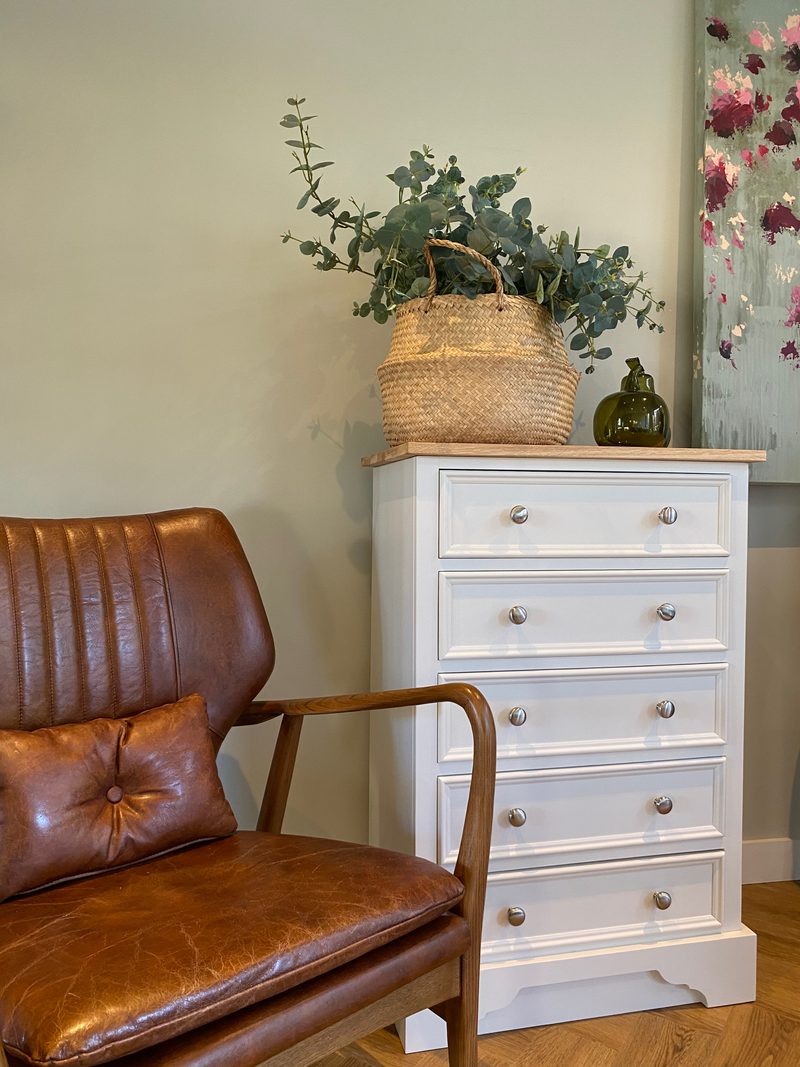 The chest photographed past a tan leather armchair, its oak top holding eucalyptus in a woven basket and a green glass bottle, with an abstract floral canvas hanging behind