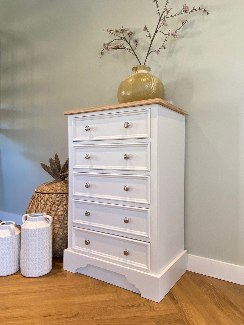 Three-quarter view of the chest showing a painted side panel, five moulded drawers and the oak top with a gold glazed vase of blossom, beside a wicker basket and two white jugs